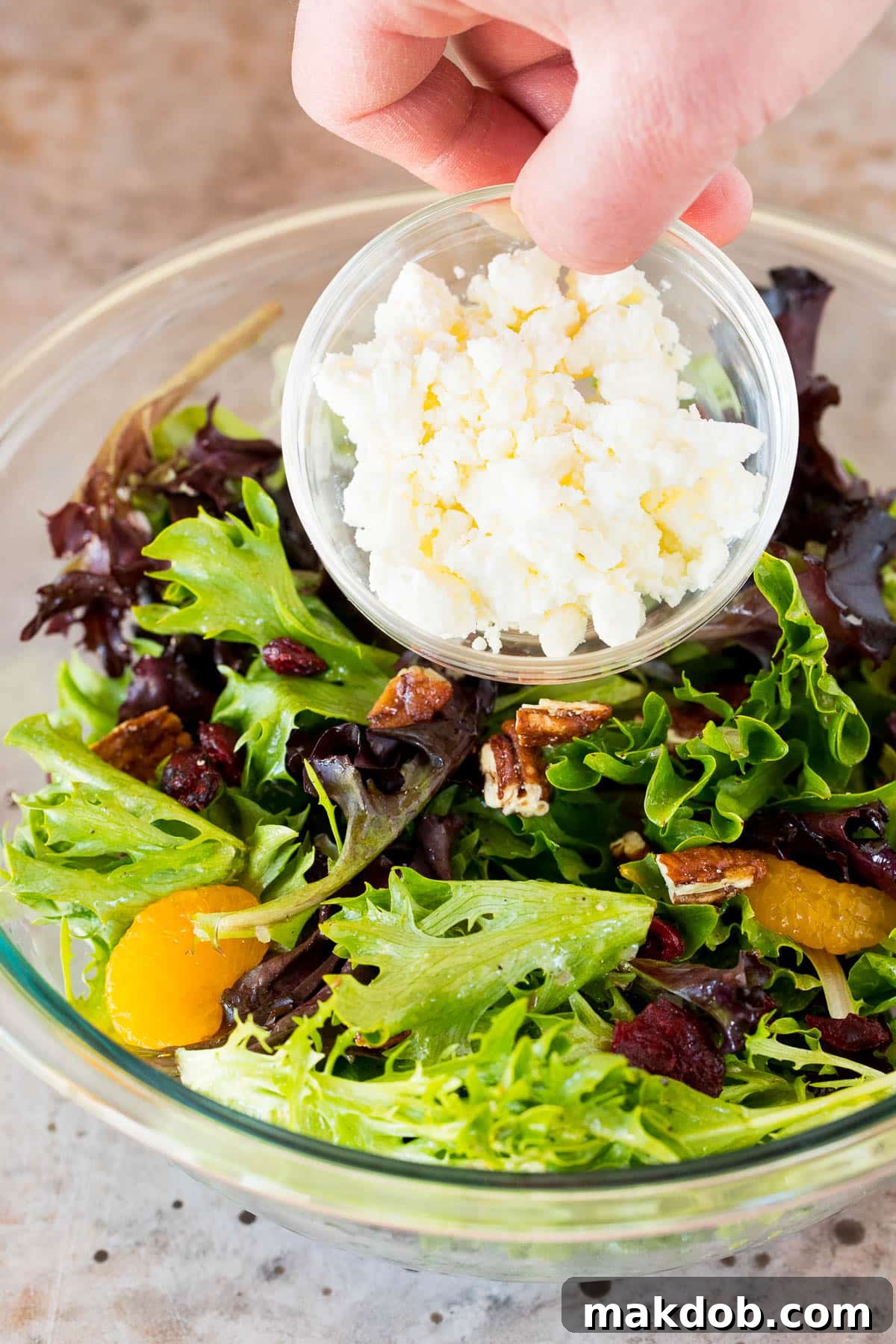 Sunshine Mandarin Salad 5 Crumbled feta cheese being poured over a fresh green salad with mandarin oranges and cranberries.