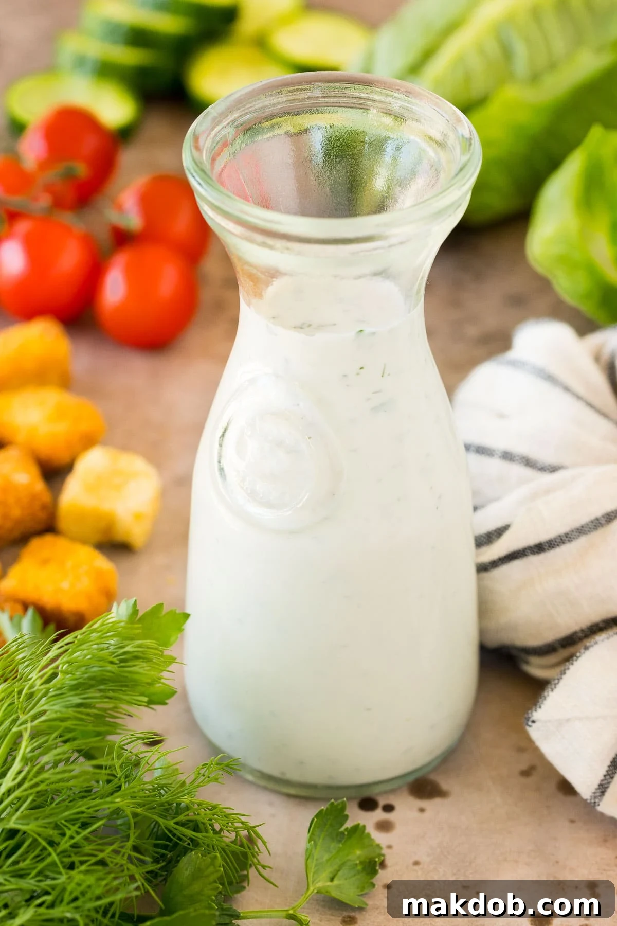 A glass container of homemade ranch dressing surrounded by fresh salad ingredients like cherry tomatoes, cucumber, and lettuce.