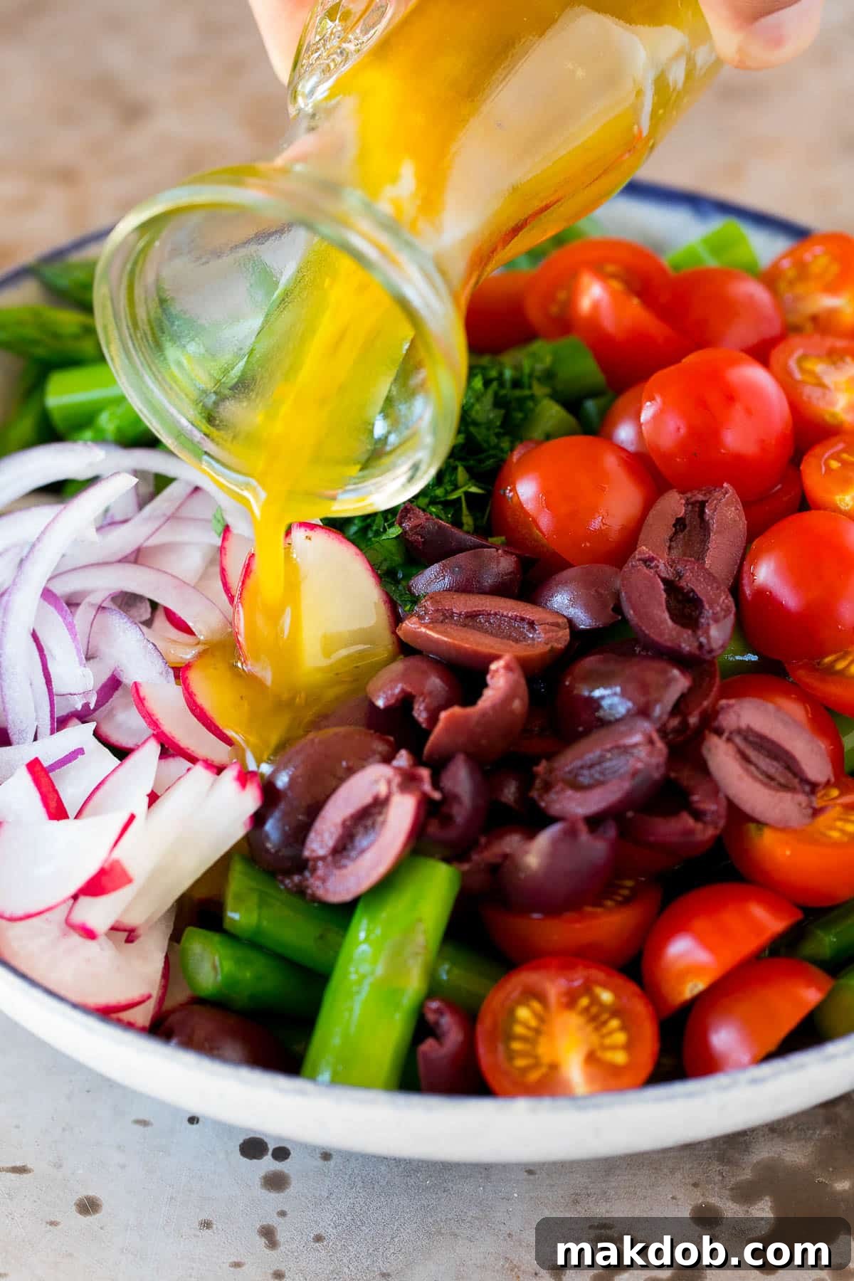 Spring Asparagus Salad 6 Homemade dressing being drizzled over a vibrant bowl of blanched asparagus, tomatoes, and olives.