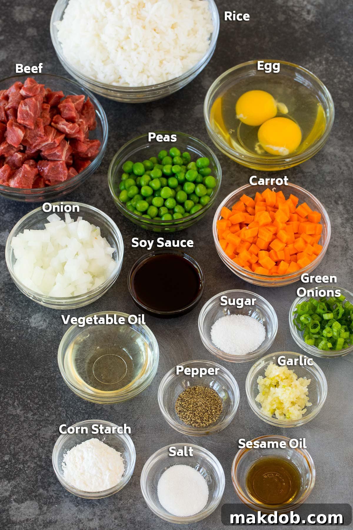Individual bowls showcasing the fresh ingredients for beef fried rice: raw steak cubes, cooked rice, assorted vegetables like carrots, peas, and onions, and a selection of seasonings and sauces.
