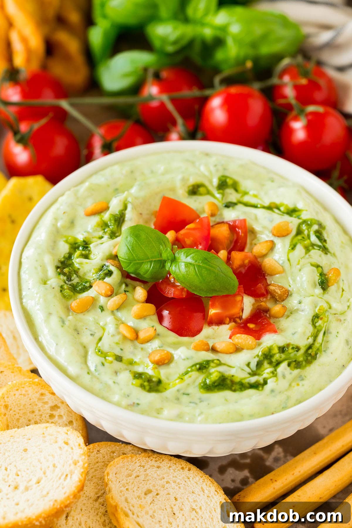 A bowl of creamy pesto dip topped with fresh diced tomatoes and basil, surrounded by an assortment of crunchy bread slices and colorful raw vegetables like carrots, celery, and bell peppers, ready for dipping.