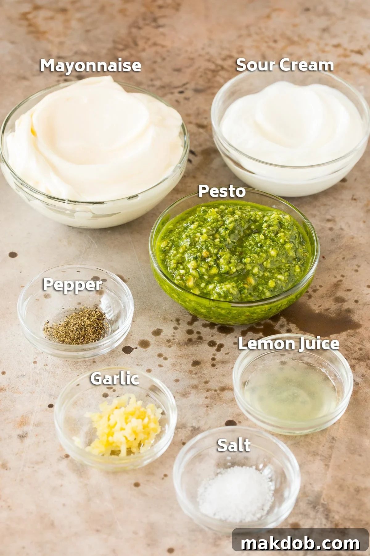 Individual bowls containing the essential ingredients for creamy pesto dip: rich mayonnaise, tangy sour cream, vibrant basil pesto, and small dishes of salt, pepper, minced garlic, and fresh lemon juice.