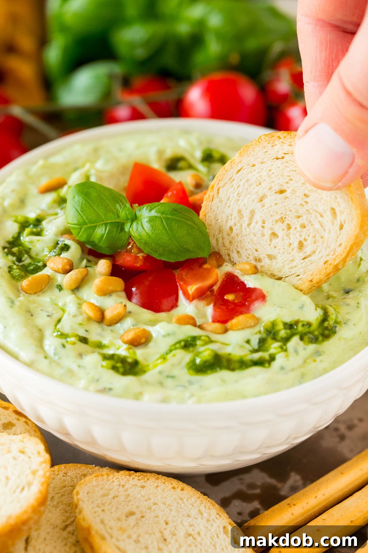 A hand gently scoops a generous portion of creamy pesto dip with a piece of artisanal bread, demonstrating the dip's rich, inviting texture and its perfect pairing with various dippers.