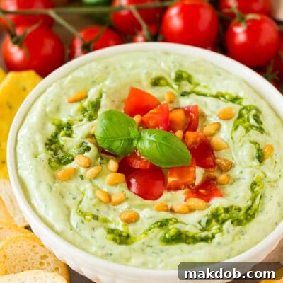 A bowl of creamy pesto dip topped with fresh diced tomatoes and basil, surrounded by an assortment of crunchy bread slices and colorful raw vegetables.
