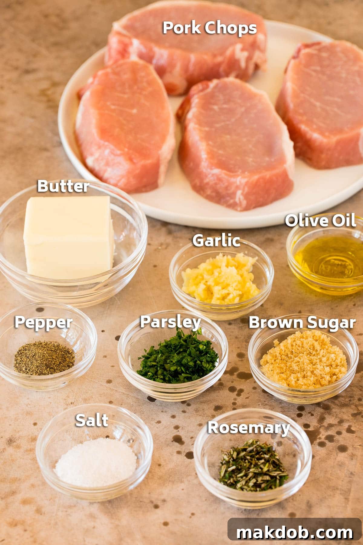 A flat lay photograph showcasing all the ingredients for baked boneless pork chops, including fresh pork chops, garlic cloves, butter, a sprig of rosemary, and various seasonings.