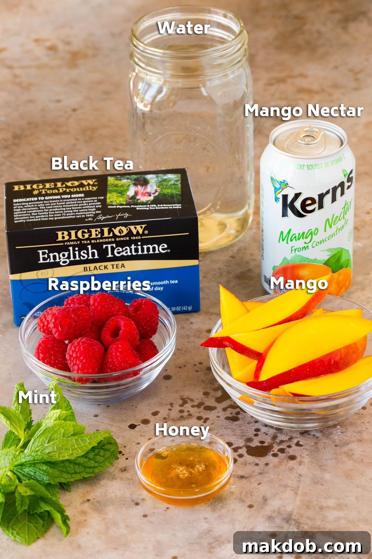 A collection of mango iced tea ingredients including black tea bags, mango nectar, fresh mango, raspberries, mint, and a honey dipper.