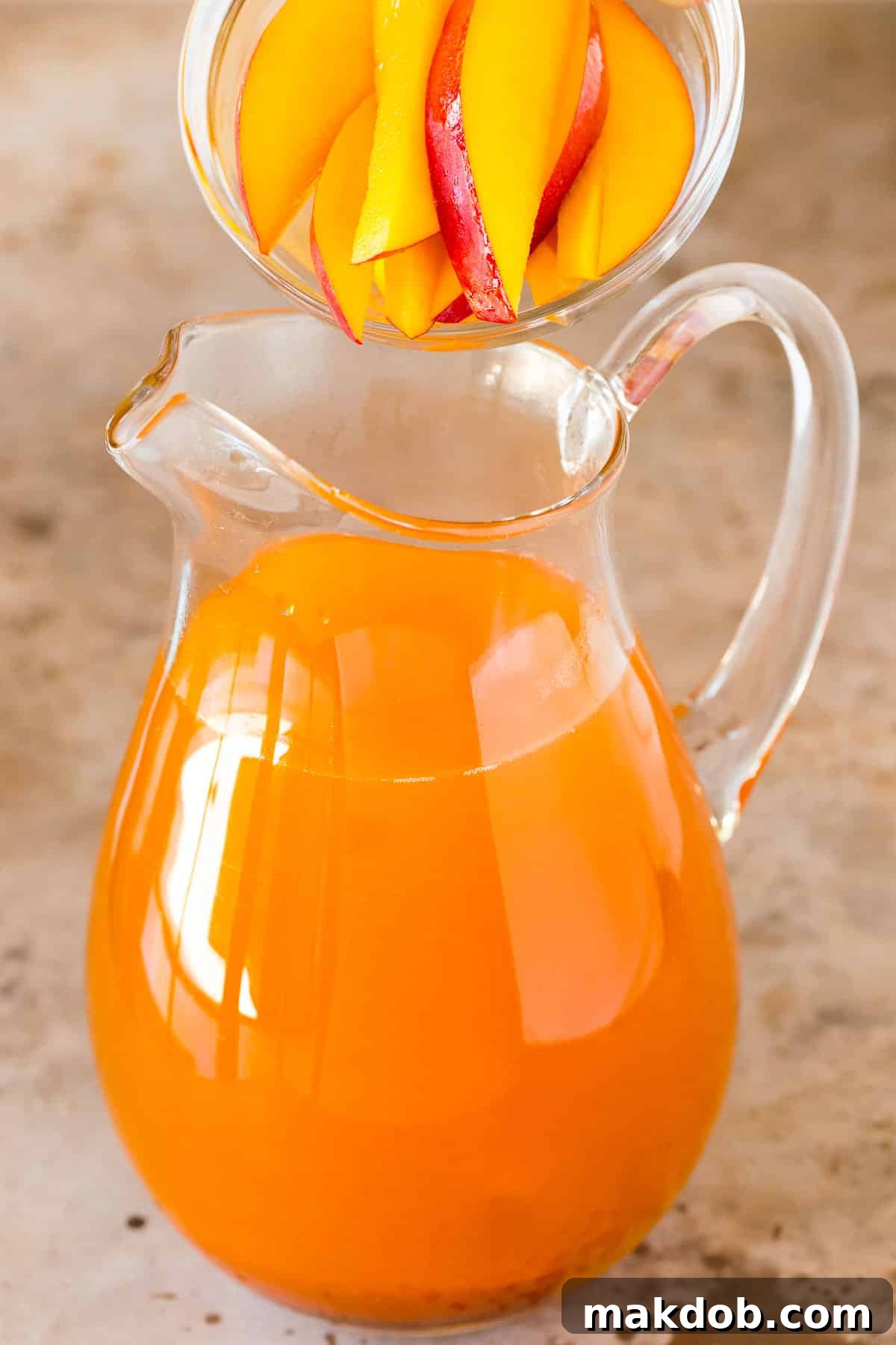 Fresh mango slices being carefully added to a large glass pitcher filled with black tea, infusing the flavor.