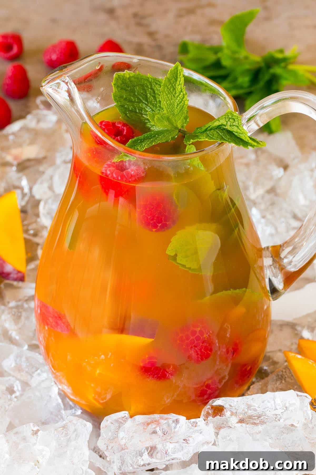 A large glass pitcher of mango iced tea filled with ice, fresh raspberries, and mint, ready to be served.