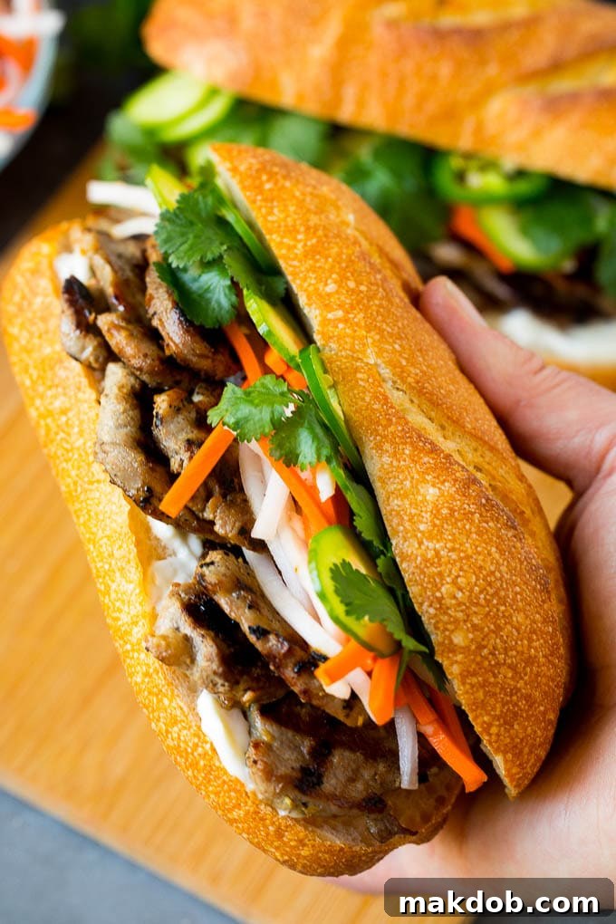 A hand presenting a perfectly constructed Banh Mi sandwich, revealing its layers of grilled pork, pickled vegetables, and fresh herbs in a crisp baguette.