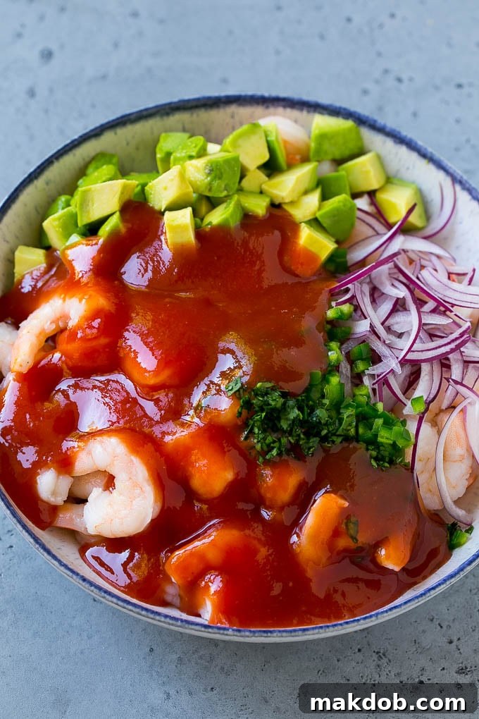 A top-down view of Mexican shrimp cocktail in a glass bowl, showcasing the cooked shrimp, diced avocado, thinly sliced red onion, and fresh cilantro, all generously coated in the vibrant red cocktail sauce.