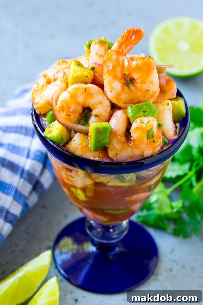An individual serving of Mexican shrimp cocktail in a clear glass, beautifully arranged with shrimp, red onion, and avocado, garnished with a lime wedge, ready to be enjoyed.