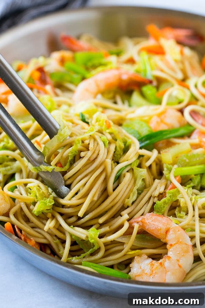 Tongs lifting a generous portion of Shrimp Chow Mein from a large pan, showcasing tender shrimp, noodles, and crisp vegetables.