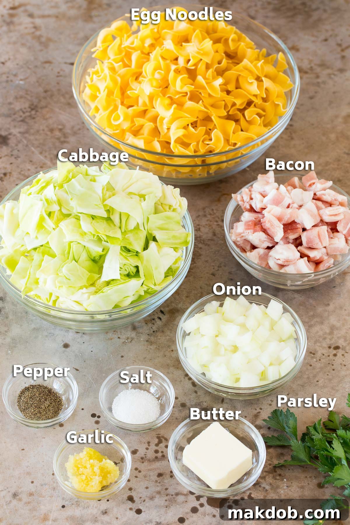 Bowls of essential Haluski ingredients including uncooked egg noodles, chopped cabbage, diced bacon, sliced onion, and garlic.