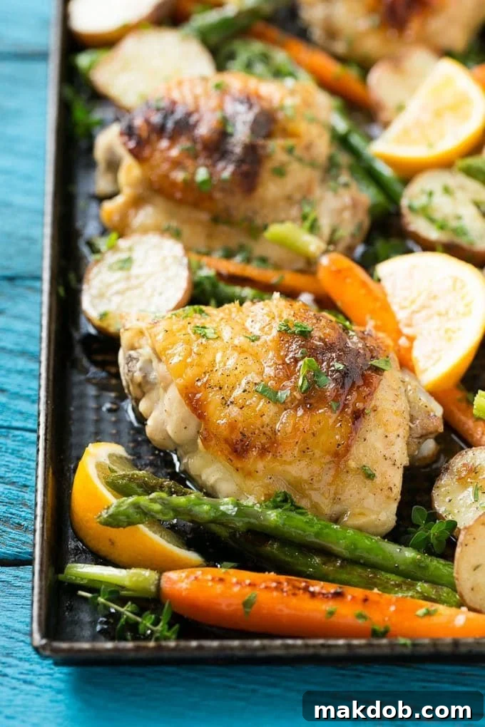 Close-up of golden brown lemon thyme chicken thighs, roasted new potatoes, and bright green asparagus on a baking sheet, seasoned with herbs.
