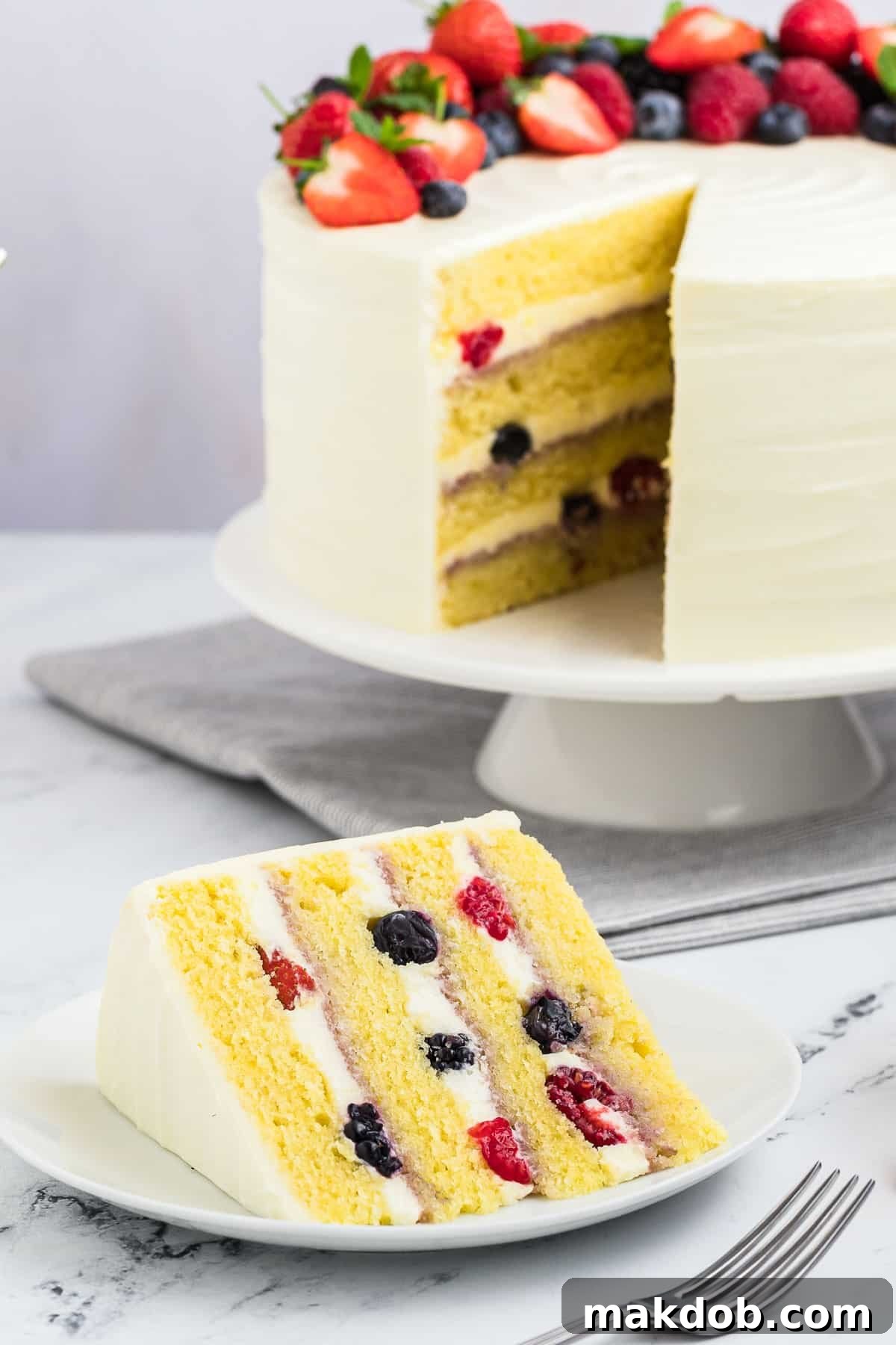 A perfectly sliced Berry Chantilly Cake resting on a white plate, showcasing its beautiful layers of yellow cake, creamy frosting, and fresh mixed berries, with a fork poised for a delicious bite.