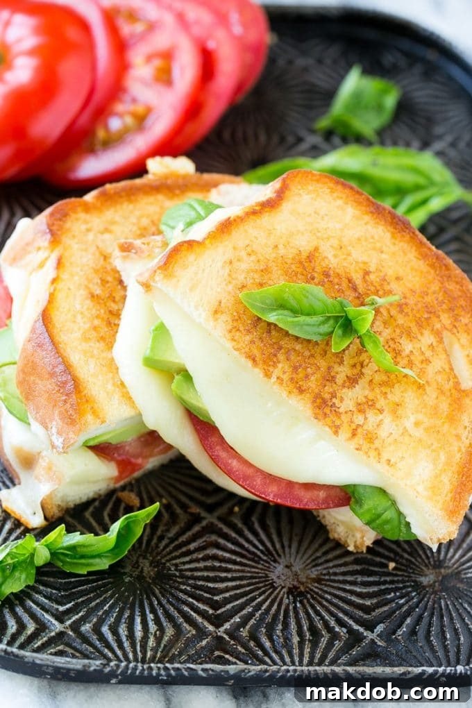 A freshly grilled Caprese sandwich, expertly cut in half to reveal layers of melted mozzarella, juicy tomatoes, fresh basil, and creamy avocado.