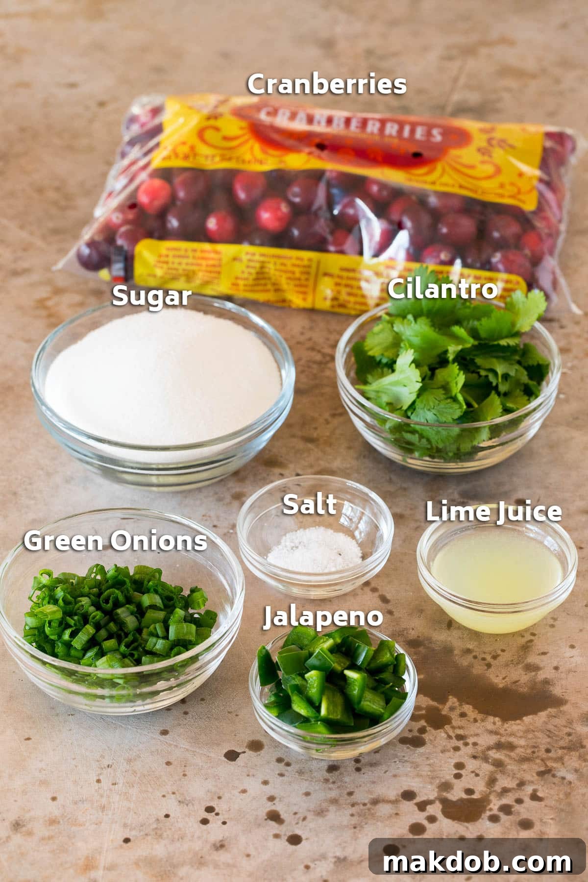 A collection of fresh ingredients laid out: bright red cranberries, vibrant lime halves, green cilantro sprigs, finely chopped green onions, and white granulated sugar.