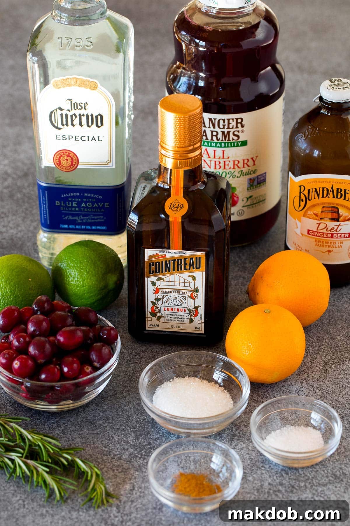 Jingle Bell Margarita 3 A selection of Christmas Margarita ingredients including bottles of tequila and triple sec, fresh oranges and limes, cranberries, and a bowl of sugar.