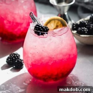 A picture of a Blackberry Bramble Cocktail, garnished with fresh blackberries and a lemon slice, on a rustic wooden table.