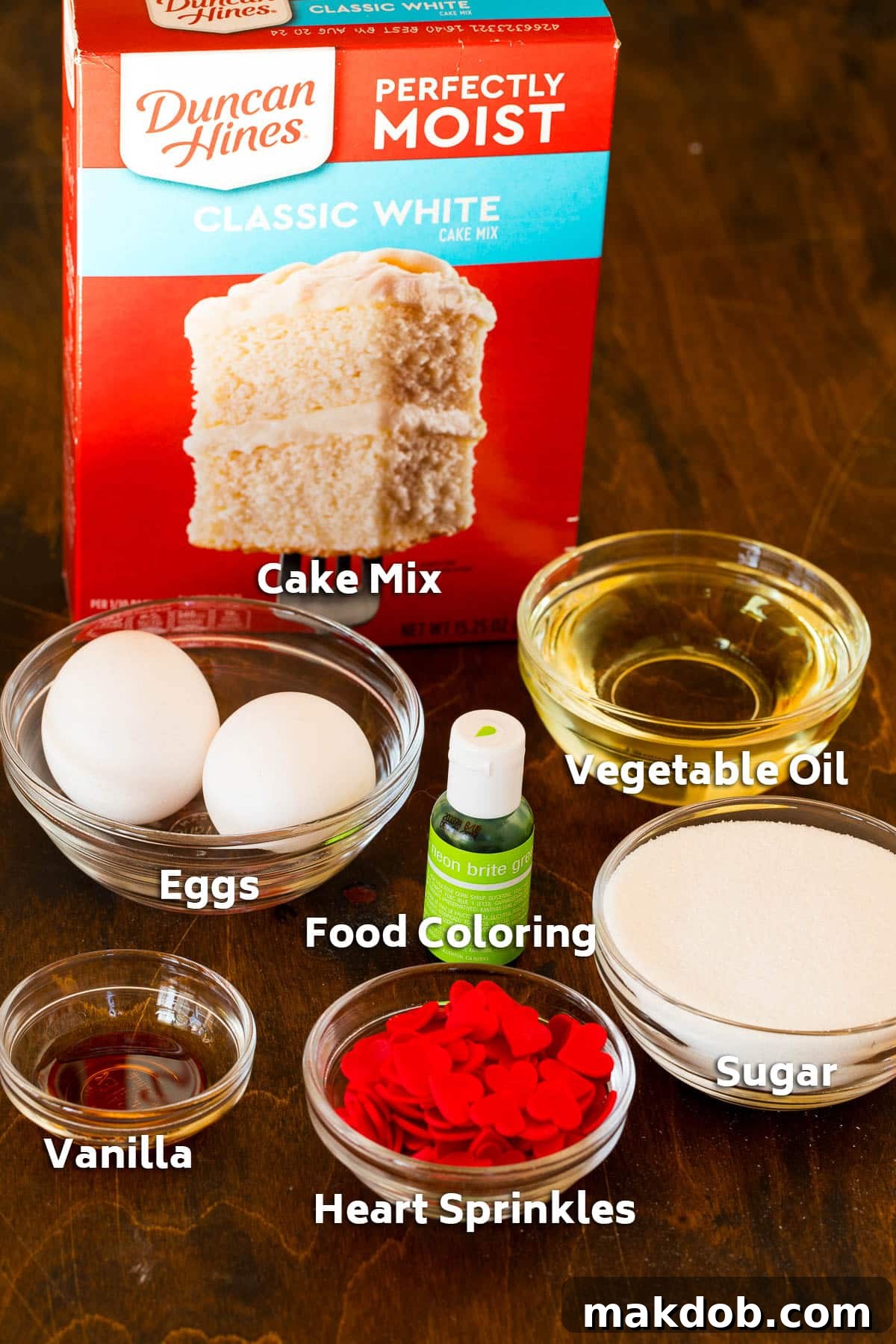 Merry Grinchmas Cookies 3 Essential ingredients for Grinch cookies laid out, including white cake mix, eggs, vegetable oil, vanilla extract, and green food coloring.