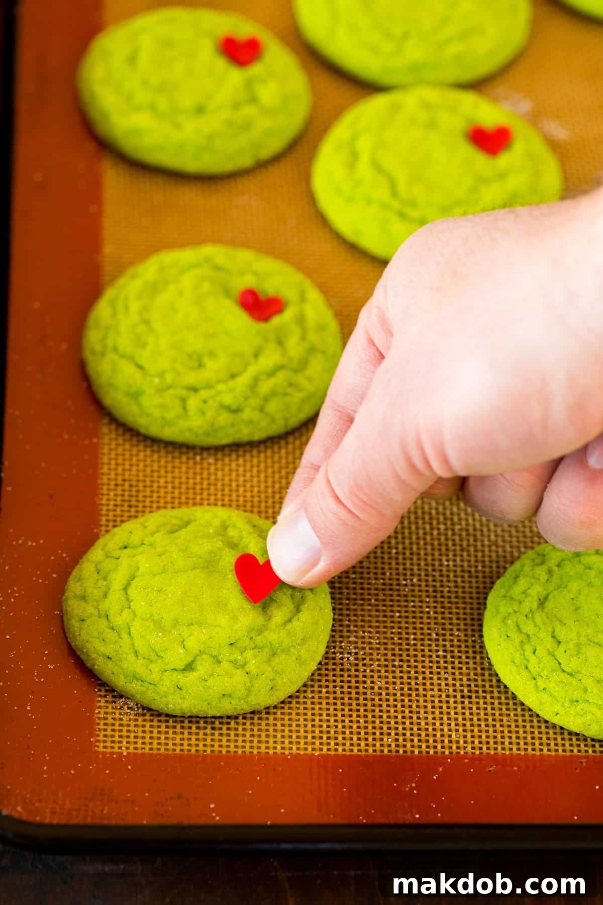 Merry Grinchmas Cookies 5 A close-up shot of a hand gently pressing a red heart sprinkle onto a freshly baked green Grinch cookie.
