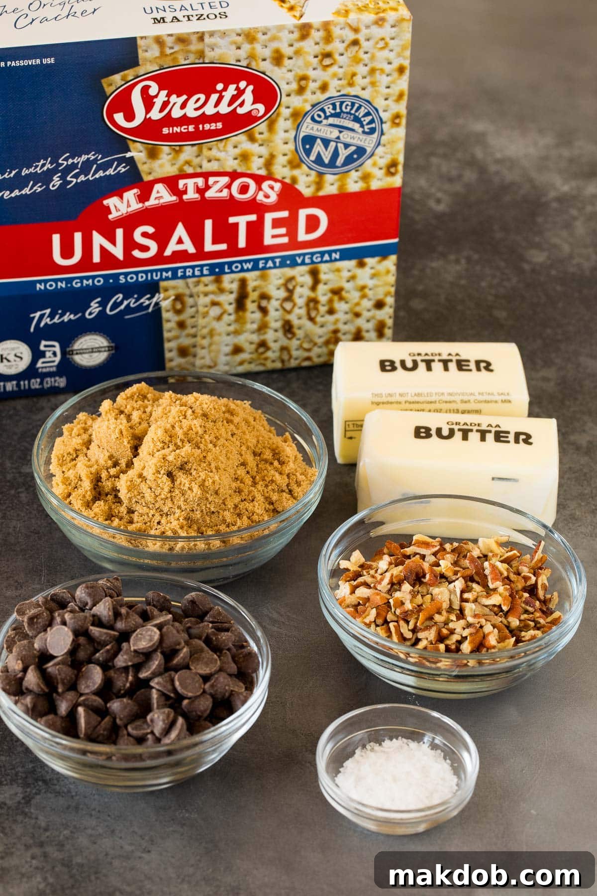 Matzo Toffee Brittle Bliss 3 Ingredients for matzo crack, including matzo, pecans, sea salt, chocolate chips, butter, and brown sugar, laid out on a white surface.