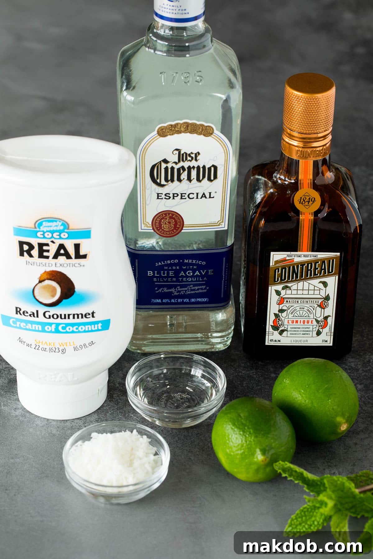 Paradise Coconut Margarita 3 A selection of fresh ingredients laid out, including cream of coconut, a bottle of triple sec, a bottle of tequila, fresh limes, and a bowl of sugar, all essential for making a delicious coconut margarita.