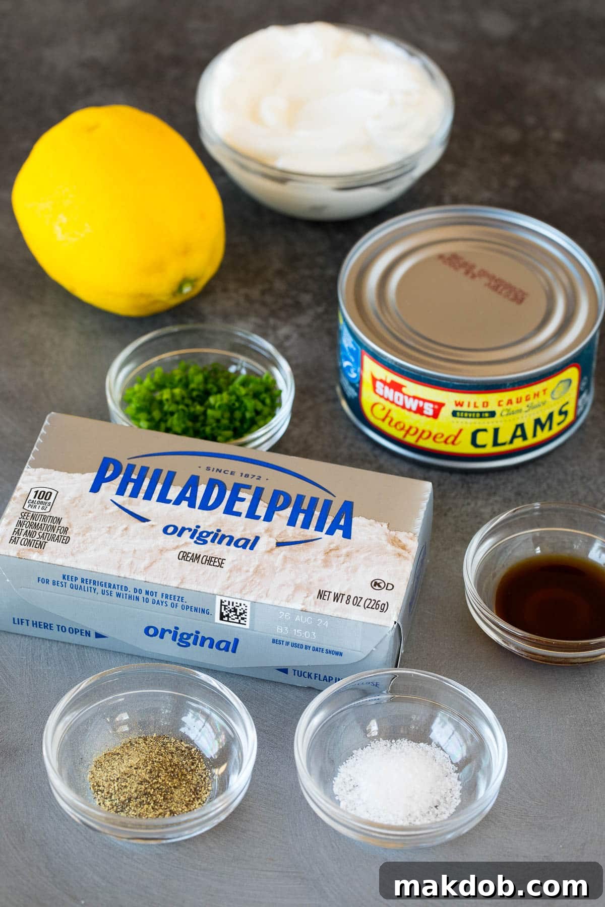 An array of fresh ingredients for clam dip, including softened cream cheese, rich sour cream, a can of chopped clams, vibrant lemon, and fresh herbs.