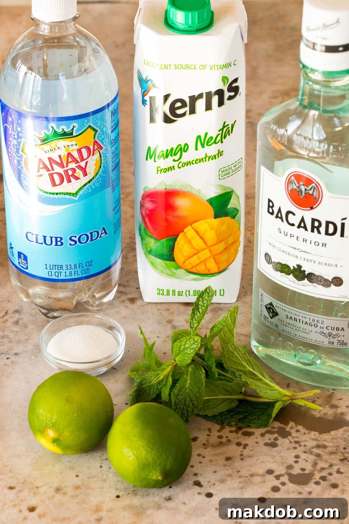 High-quality bottles of white rum, mango nectar, and club soda arranged with fresh limes, granulated sugar, and mint leaves, ready for cocktail preparation.