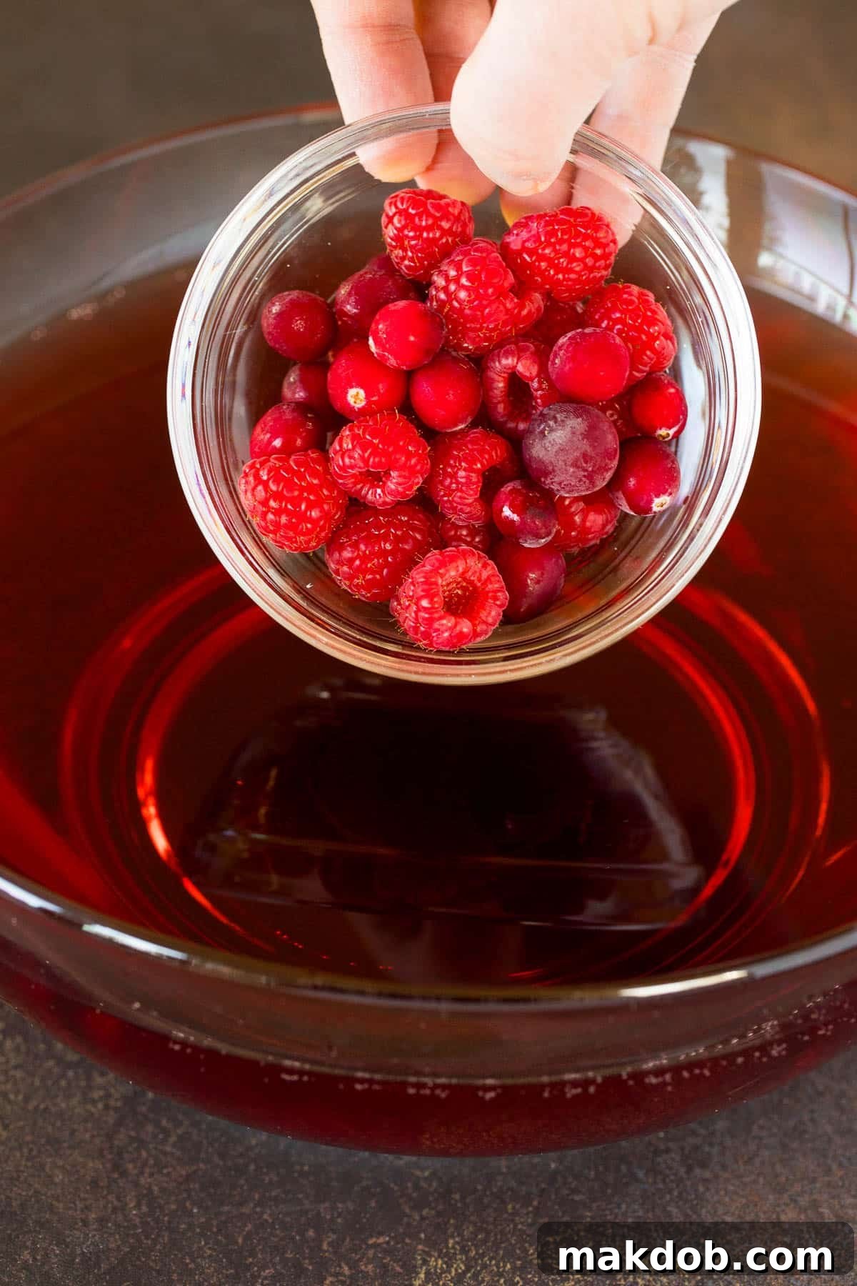Fresh raspberries and cranberries being carefully poured into a festive bowl of Halloween punch.