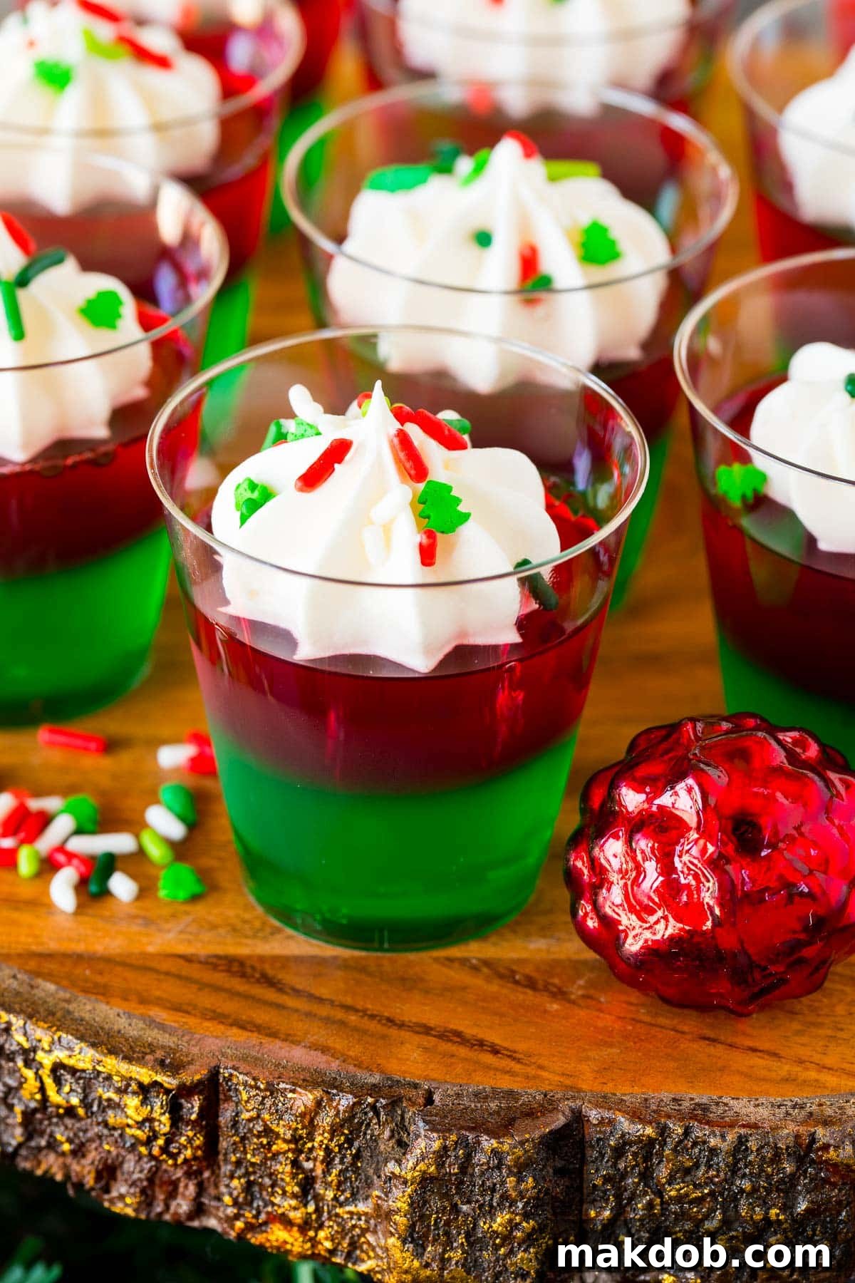 Beautifully layered Christmas Jello shots in clear cups, garnished with whipped cream and festive red and green sprinkles, ready for a holiday party.