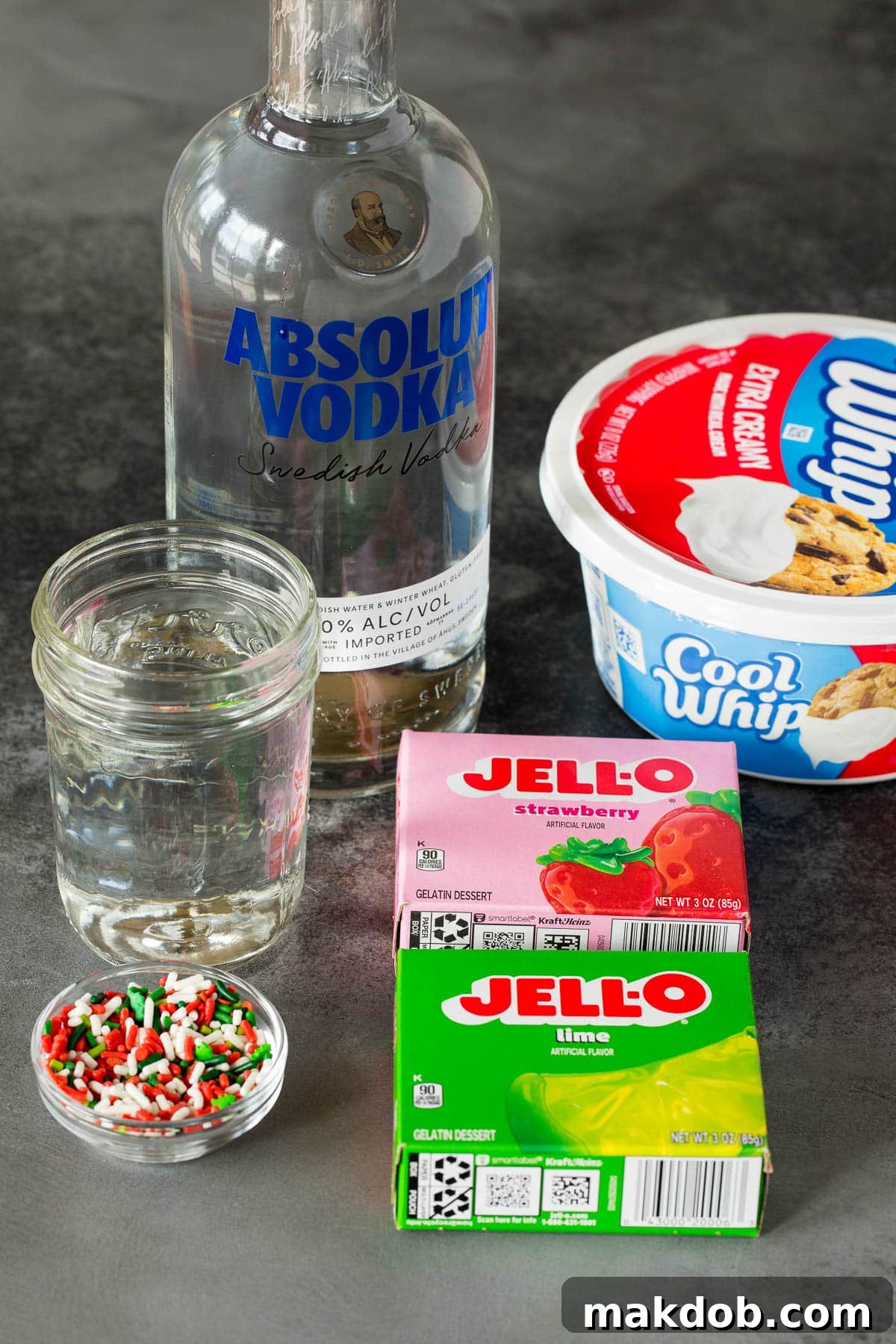 A flat lay photograph showcasing the key ingredients for Christmas Jello shots, including boxes of red and green Jello, a bottle of vodka, a measuring cup with water, and colorful holiday sprinkles.