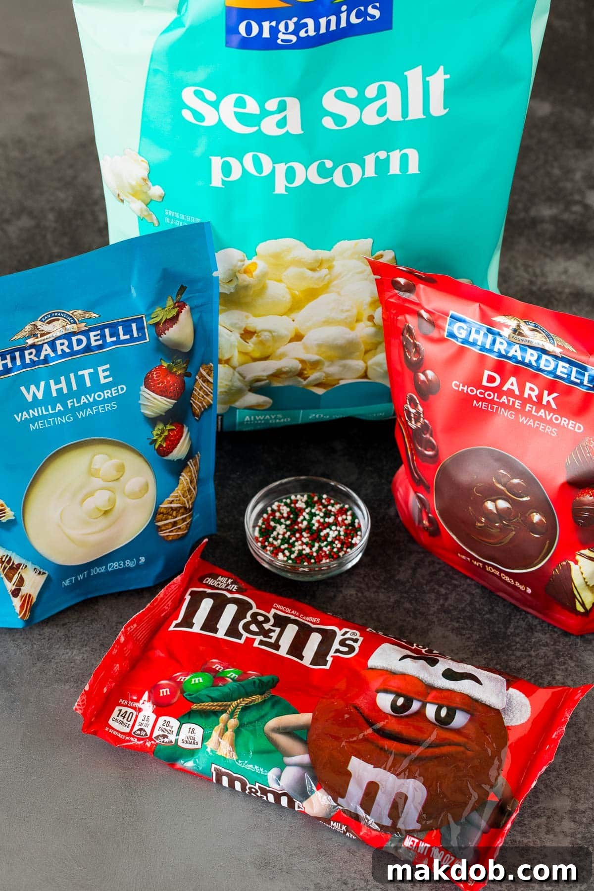 Sparkling Holiday Popcorn 3 Ingredients for Christmas popcorn, including plain popcorn, white and dark candy melts, Christmas M&M's, and festive sprinkles, laid out on a white surface.