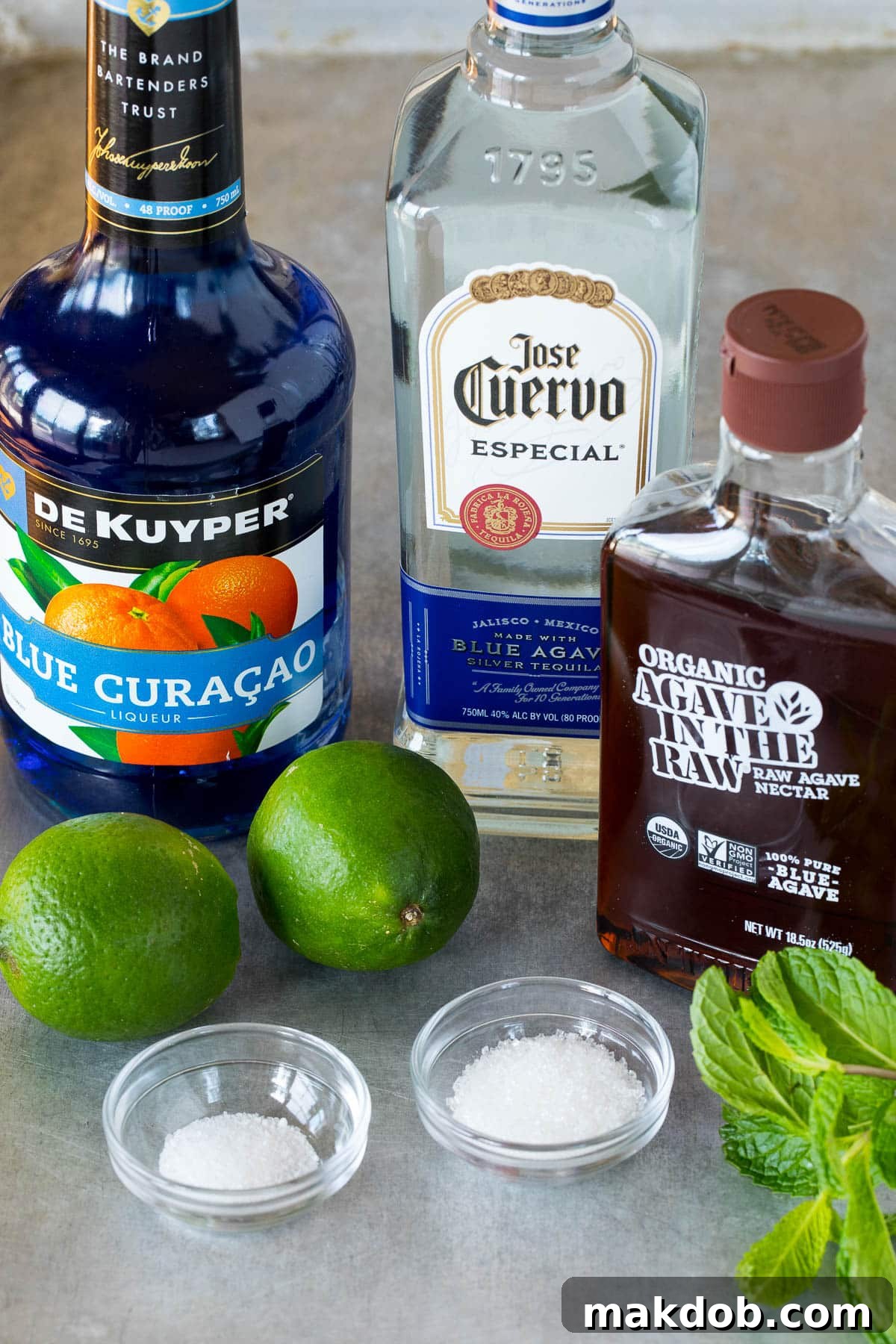 Electric Blue Margarita 3 Ingredients including bottles of liquor, limes, mint and sugar.