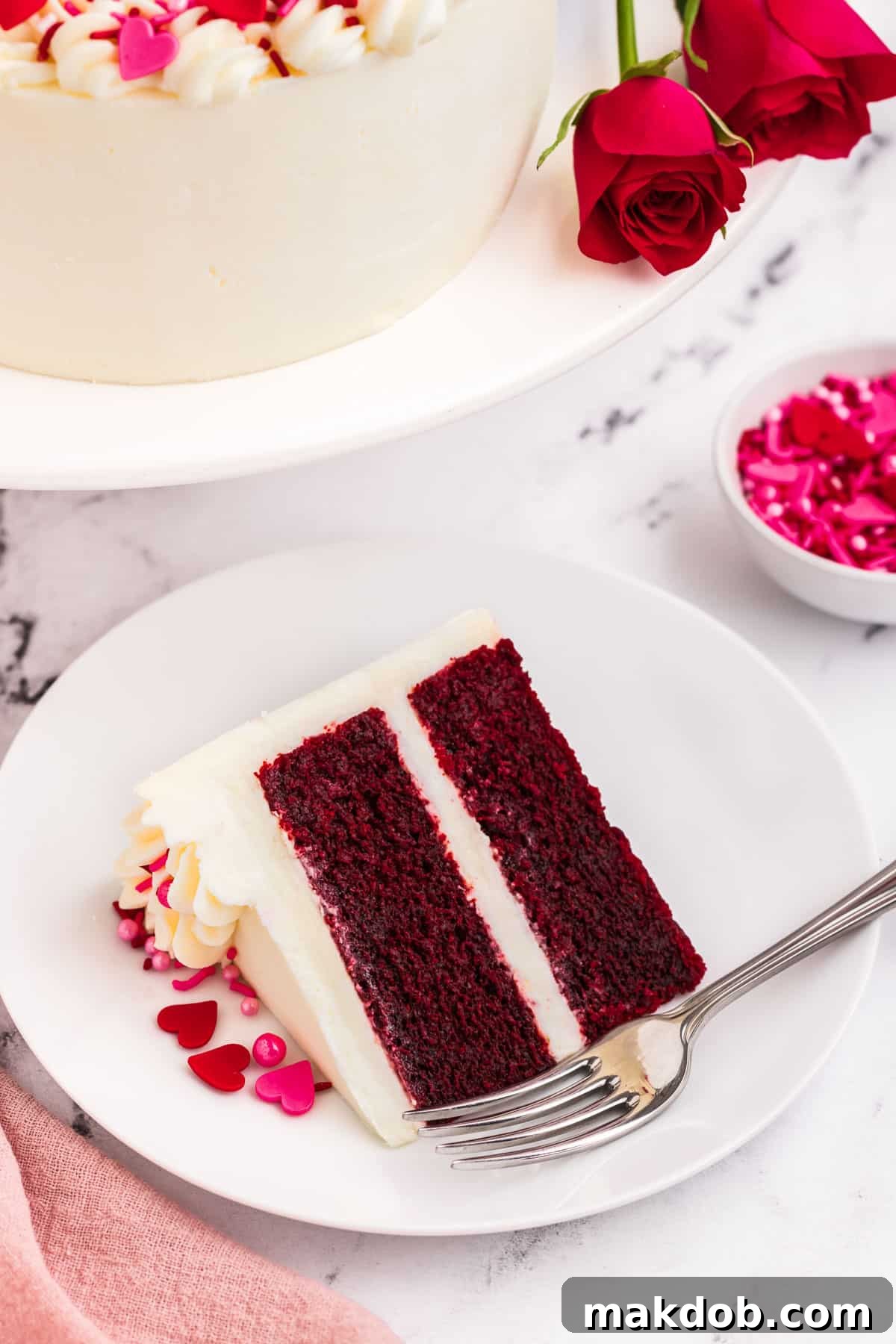 A perfectly sliced piece of red velvet heart cake, showing its vibrant red layers and creamy white frosting, served on a plate with a fork.