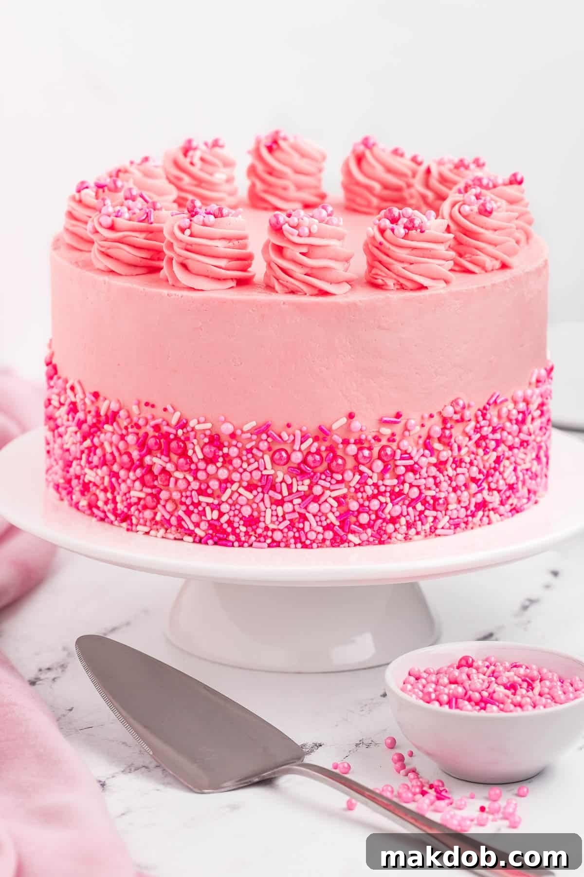 A beautifully decorated Pink Velvet Cake on a white pedestal, adorned with sprinkles and topped with piped rosettes of frosting.