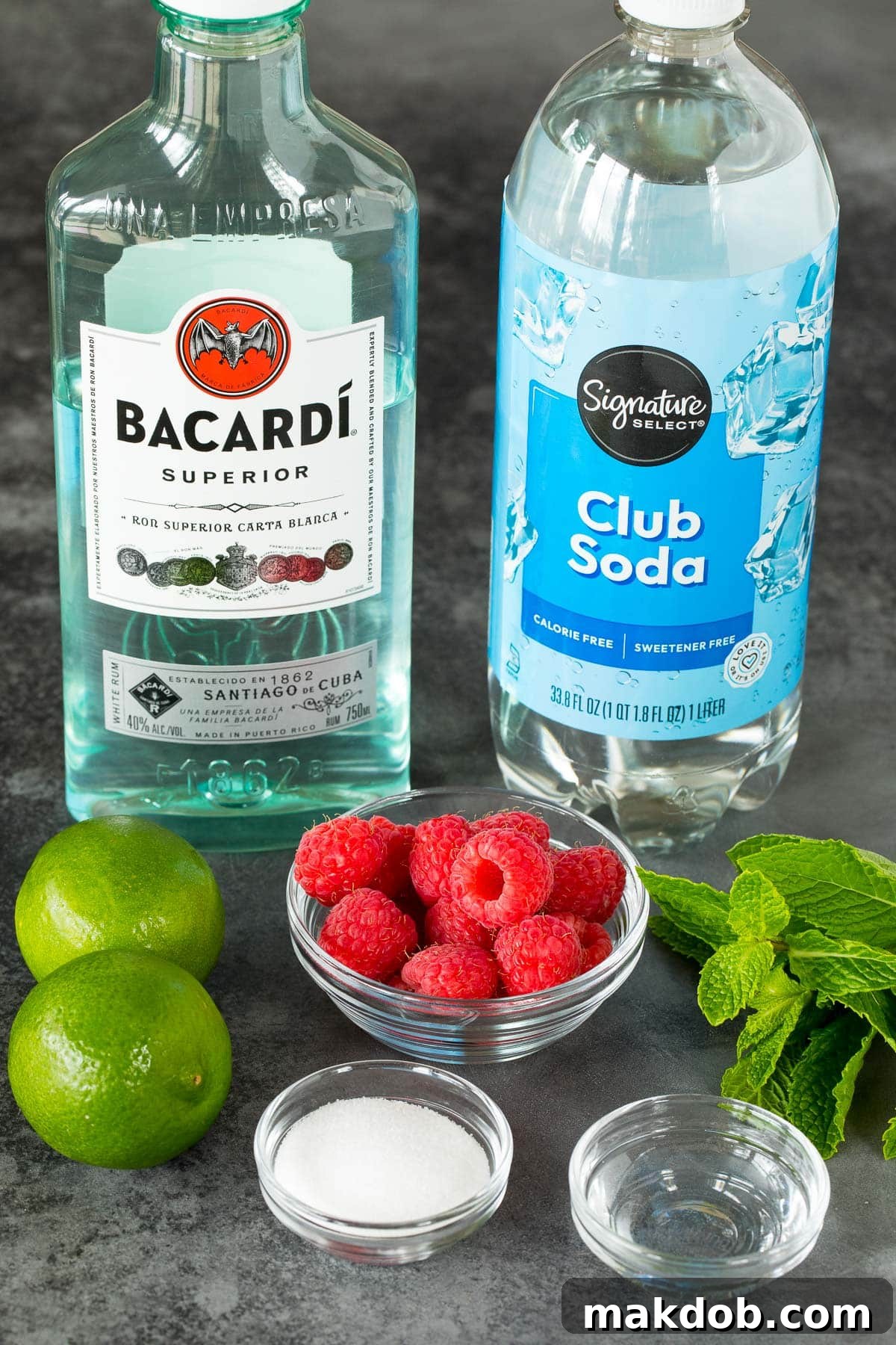 Key ingredients for a Raspberry Mojito laid out: fresh limes, mint leaves, granulated sugar, white rum, club soda, and ripe raspberries.