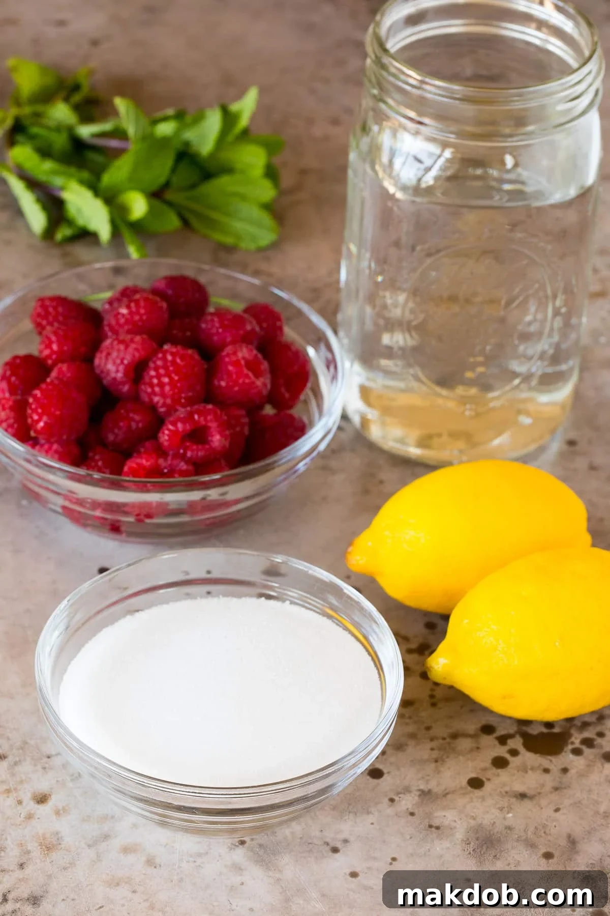 A flat lay of fresh ingredients laid out on a white surface, including bright red raspberries, whole lemons, granulated sugar, a glass of water, and a sprig of fresh mint, indicating what is needed for the lemonade.