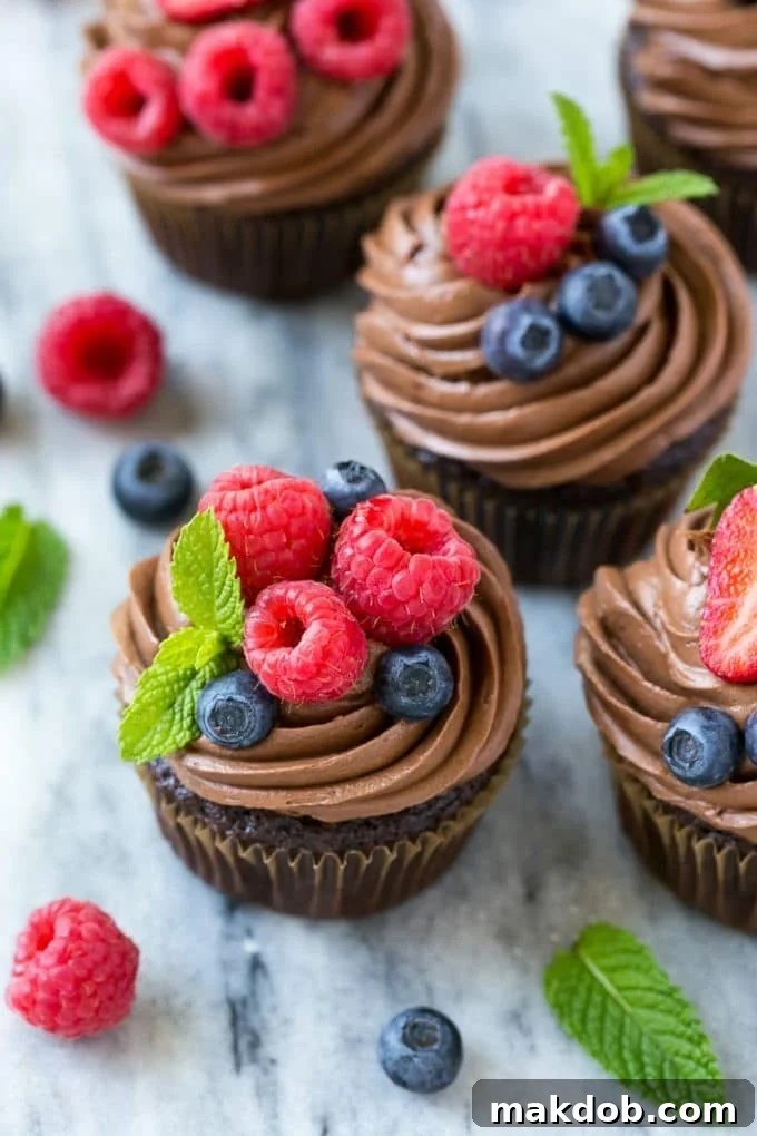 Close-up of three chocolate hazelnut cupcakes, beautifully frosted and garnished with raspberries, blueberries, and fresh mint. The rich chocolate and smooth hazelnut flavors are a perfect pairing.