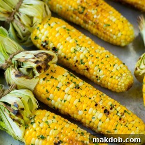 Perfectly grilled corn on the cob resting on a sheet pan, each ear beautifully charred and glistening with melted garlic and herb butter. The vibrant green of the herbs adds a fresh touch, indicating a delicious and aromatic flavor that's ideal for any outdoor gathering or summer meal.