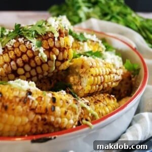 A close-up of spicy ancho grilled corn on the cob, richly seasoned with a reddish chili blend. The corn is artfully paired with a creamy dollop of green avocado crema and sprinkled with white cotija cheese, promising a harmonious blend of smoky, spicy, and cooling flavors for an irresistible gourmet experience.