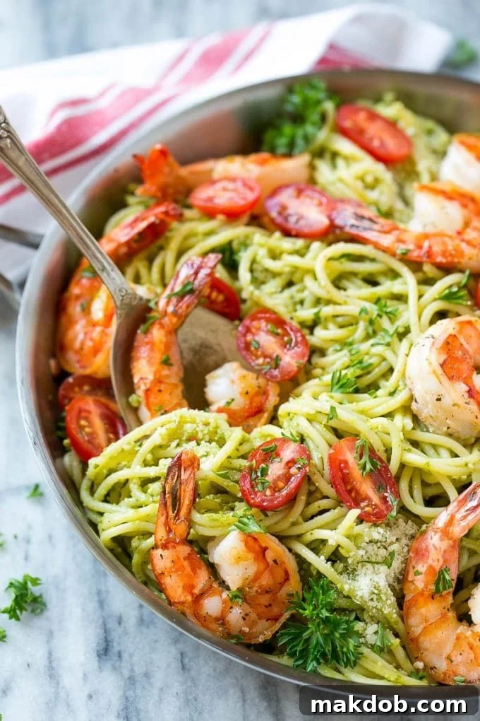 A serving spoon lifting a portion of shrimp pesto pasta from a pan, highlighting the perfectly coated spaghetti, plump shrimp, and vibrant tomatoes.