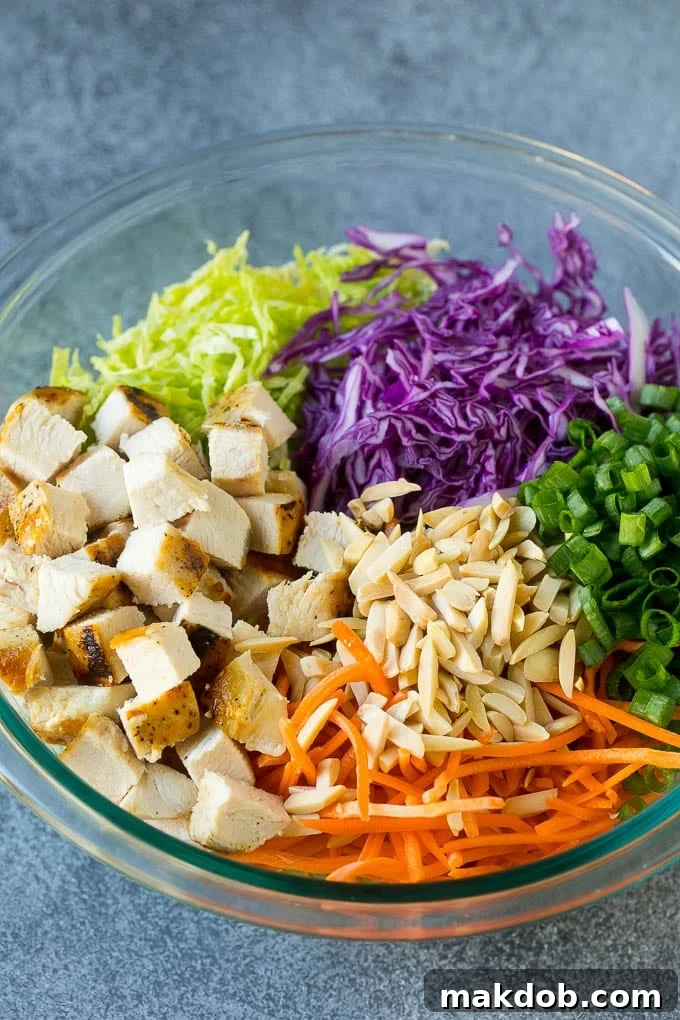 A large bowl filled with vibrant green and purple cabbage, shredded carrots, tender chicken, and crisp green onions, awaiting the dressing.