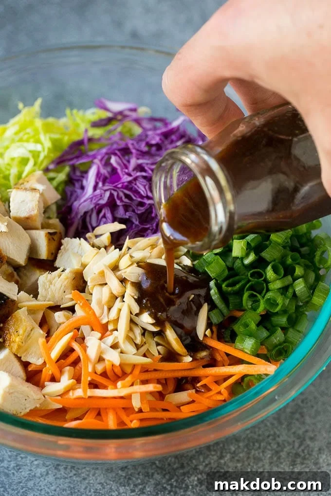 A close-up shot of the sesame ginger dressing being generously poured over a colorful mix of cabbage, carrots, chicken, and green onions in a large bowl.