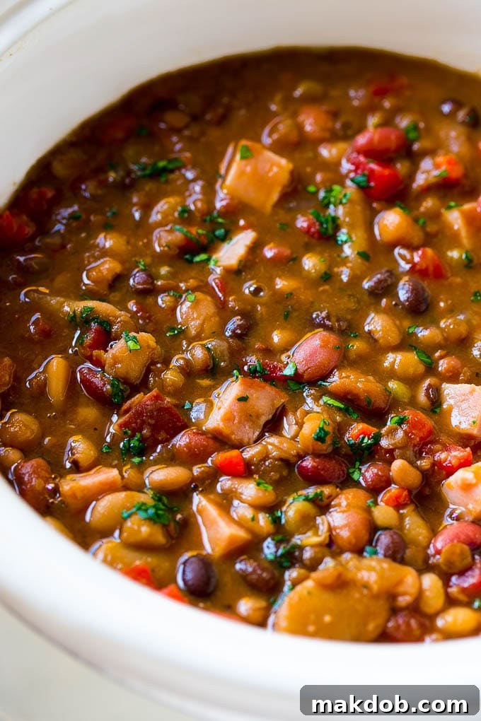 Slow Cooker Ham Bone Soup | Ham and Bean Soup | Ham Bone and Bean Soup | Ham Soup | Leftover Ham Recipe #ham #hamsoup #leftoverham #soup #slowcooker #crockpot #dinneratthezoo A close-up shot of a slow cooker filled with simmering ham bone soup, richly colored with a blend of beans, carrots, and other vegetables, garnished with fresh parsley on top.