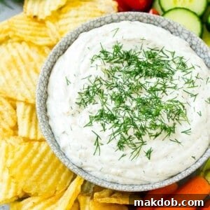 A bowl of creamy dill dip surrounded by potato chips and fresh vegetables, perfect for snacking.