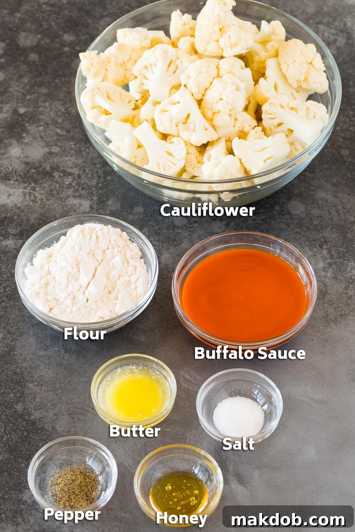 Bowls of ingredients including fresh cauliflower florets, a bottle of buffalo wing sauce, and various seasonings, ready for preparation.