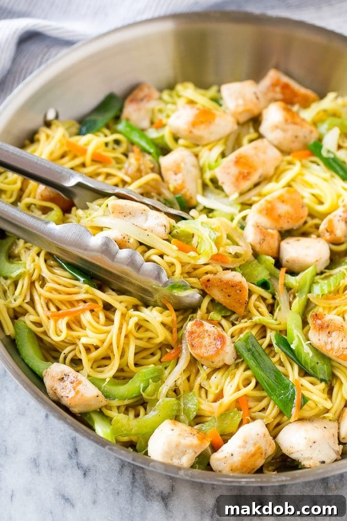 A large pan filled with vibrant chicken chow mein, ready to be served, with serving tongs resting in the dish.