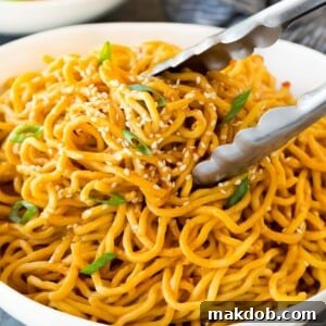 A pair of tongs gracefully serving up a portion of glossy sesame noodles, glistening with a rich, savory peanut and sesame sauce, ready to be enjoyed.