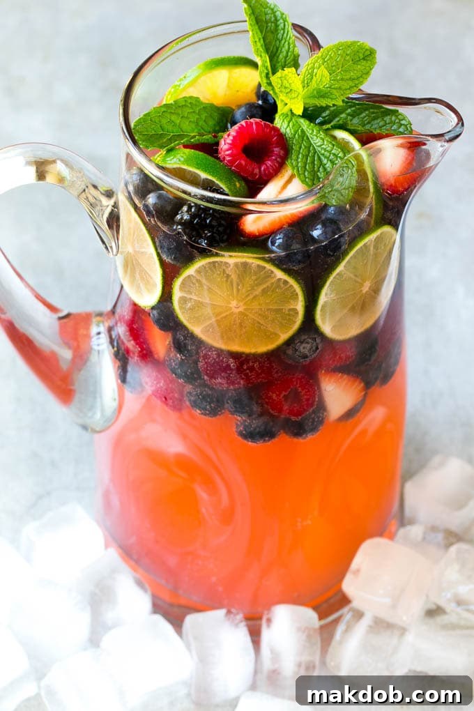 A large, clear pitcher filled with a vibrant pink cocktail, generously garnished with various fresh fruits and mint leaves.